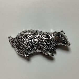 Może przedstawiać: Srebrna figurka borsuka. Borsuk jest przedstawiony w pozycji chodu z głową zwróconą w bok. Figurka jest szczegółowo wykonana z fakturowanym futrem.