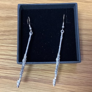 May include: A pair of silver wand earrings in a black box. The earrings are long and slender, with a detailed design that resembles a wand.