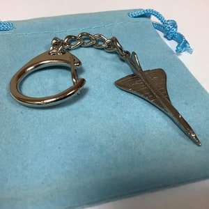 May include: A silver keychain featuring a detailed model of a Concorde supersonic jet. The keychain is attached to a silver ring and chain, and comes in a light blue velvet pouch.