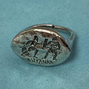 May include: A silver ring with a raised design of three figures holding swords. The ring is on a light blue velvet pouch. The inscription "ASTYANAX" is visible on the ring.