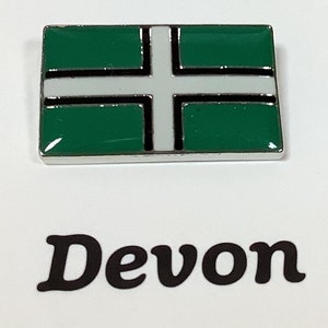 Könnte beinhalten: Eine silberne Anstecknadel mit einem grünen, weißen und schwarzen Flaggen-Design. Die Flagge hat ein weißes Kreuz auf grünem Hintergrund mit schwarzen Rändern. Der Text "Devon" ist unter der Flagge gedruckt.