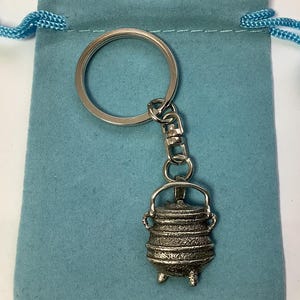 May include: A silver pot keychain with a ring and chain. The pot is detailed with a textured surface and has three legs. The keychain is displayed on a blue velvet pouch.