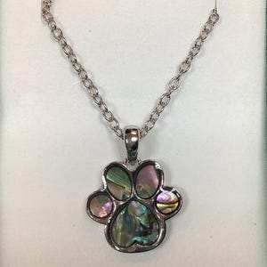 May include: A silver-toned necklace featuring a paw print pendant. The pendant has a silver border and is filled with iridescent abalone shell, displaying shades of green, pink, and purple. The chain is a simple link style.