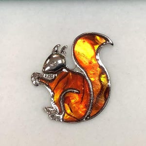 May include: A silver-toned squirrel pin with a vibrant orange and yellow enamel design. The squirrel is depicted in a sitting position, with a detailed head and tail. The pin is likely made of metal and is suitable for a jacket, bag, or hat.