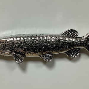 Op de afbeelding: Een zilverkleurig metalen visbeeldje, dat lijkt op een snoek of een muskellunge, met gedetailleerde schubben en vinnen.