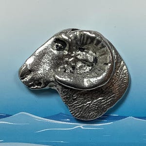 Peut inclure: Sculpture en étain argenté d'une tête de mouton avec une corne recourbée. La sculpture est détaillée avec une fourrure texturée et une expression réaliste.