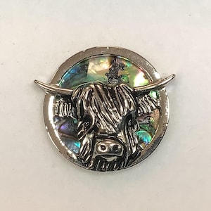May include: A silver-toned pin featuring a detailed, three-dimensional image of a Highland cow's head. The cow's fur is textured and the background is a shimmering iridescent abalone shell.