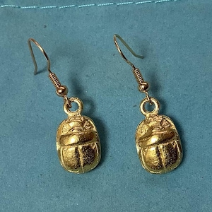 Puede incluir: Un par de pendientes de tono dorado con un diseño de escarabajo. Los pendientes están hechos de metal y tienen un cierre de gancho simple.
