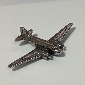 Può includere: Un modellino di aereo in argento con un emblema rotondo sulla coda e un'elica nella parte anteriore.