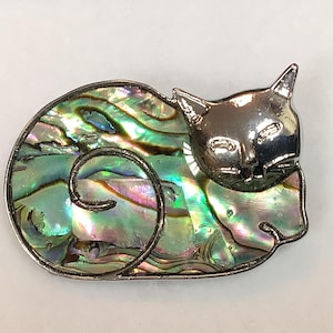 Op de afbeelding: Een zilveren broche met een slapende kat van iriserende abalone schelp.