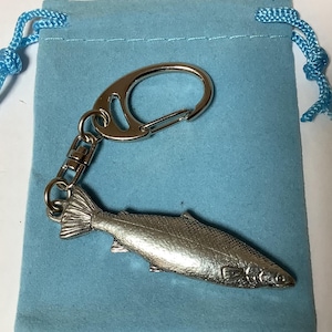 May include: A silver-colored fish-shaped keychain with a lobster clasp and a small chain, resting on a light blue drawstring pouch. The fish has detailed scales and fins. The pouch has a blue drawstring.