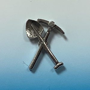 May include: A silver-toned metal charm featuring a crossed pickaxe and shovel. The tools are detailed with a textured surface.