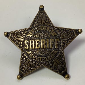 Puede incluir: Una insignia de estrella de sheriff de metal dorado con detalles intrincados. La insignia presenta las palabras "Lincoln County Sheriff" en un diseño circular.