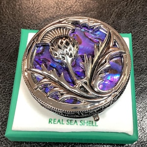 May include: A silver and purple abalone shell trinket box with a detailed design of a thistle flower. The box is in a green box with the words "REAL SEA SHELL" printed on the bottom.
