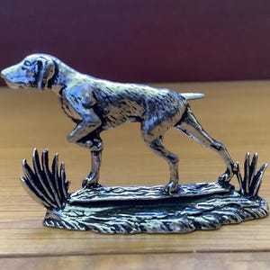 Op de afbeelding: Een zilveren beeldje van een jachthond dat op een voetstuk met gras staat. De hond staat in een rennende houding met zijn hoofd naar de zijkant gedraaid.