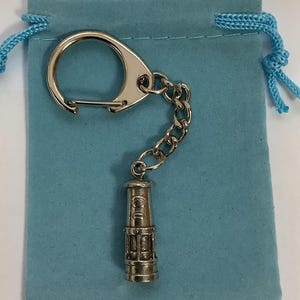 Könnte beinhalten: Ein silberner Schlüsselanhänger mit einer kleinen, detaillierten Nachbildung einer Grubenlampe. Der Schlüsselanhänger ist an einem silbernen Ring und einer Kette befestigt. Die Lampe ist etwa 5 cm hoch und hat eine zylindrische Form. Der Schlüsselanhänger wird auf einem hellblauen Samtbeutel präsentiert.