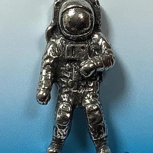 May include: A silver pewter figurine of an astronaut wearing a spacesuit with a helmet. The astronaut is standing with its arms at its sides.