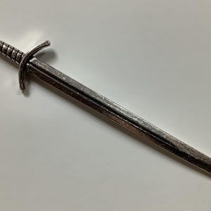 Könnte beinhalten: Ein silberfarbenes Schwert mit einem detaillierten Griff und einer spitzen Spitze. Das Schwert ist etwa 25 cm lang.