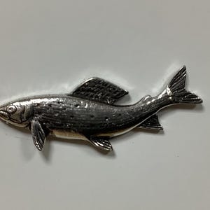 Puede incluir: Un pin de pescado de color plateado con un diseño detallado. El pez tiene un cuerpo ligeramente curvado y una aleta en la espalda.