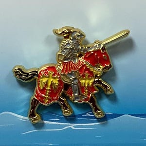 Puede incluir: Un pin de metal dorado que representa a un caballero a caballo. El caballero lleva armadura y sostiene una lanza. El caballo es rojo con detalles dorados. El pin está colocado sobre un fondo azul y blanco.