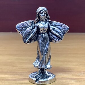 Op de afbeelding: Een zilveren beeldje van een fee met vleugels, die een lange jurk draagt en op een ronde basis staat.