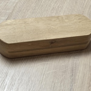 May include: A rectangular wooden box with rounded edges. The box is made of light-colored wood, possibly oak, and has a natural grain pattern. The box is closed and sits on a light-colored wooden surface.