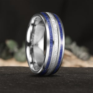 Könnte beinhalten: Ein silberner Wolframcarbid-Ring mit zwei blauen Lapislazuli-Einlegebändern. Der Ring hat eine gebürstete Oberfläche und ein Meteoritmuster.