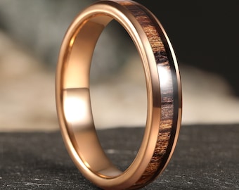 Puede incluir: Un anillo de carburo de tungsteno de color oro rosa con una incrustación de madera marrón. El anillo tiene un borde biselado y un acabado pulido.