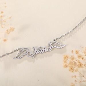 Puede incluir: Collar de plata con el nombre "Emma" escrito en cursiva con alas de &aacute;ngel a cada lado.