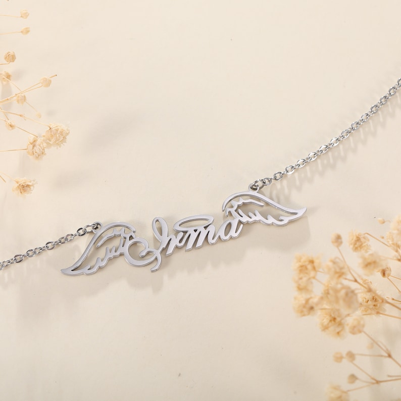 Puede incluir: Collar de plata con el nombre "Emma" escrito en cursiva con alas de &aacute;ngel a cada lado.