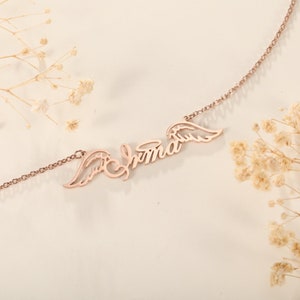 Puede incluir: Un collar de oro rosa con el nombre "Emma" y alas de &aacute;ngel a cada lado.