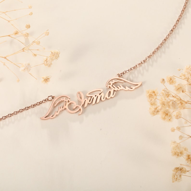 Puede incluir: Un collar de oro rosa con el nombre "Emma" y alas de &aacute;ngel a cada lado.