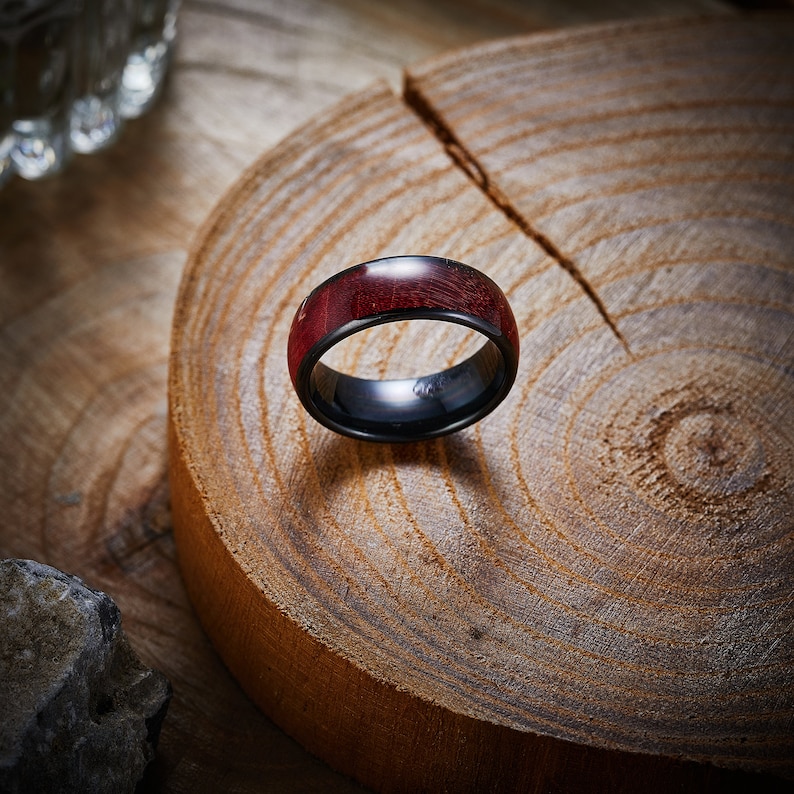 May include: A black and red ring with a wood inlay. The ring is sitting on a wooden surface.