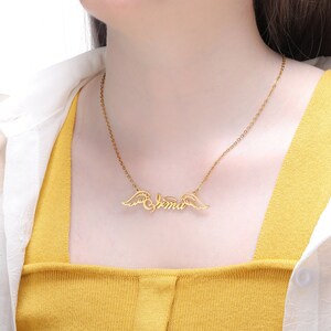 Puede incluir: Collar de oro con una cadena delicada y un colgante con el nombre "Emma" con alas de &aacute;ngel a cada lado.