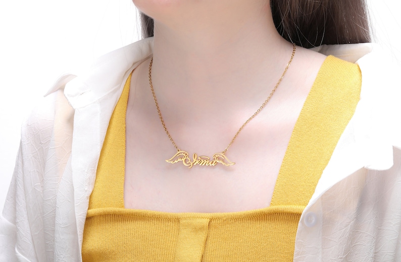 Puede incluir: Collar de oro con una cadena delicada y un colgante con el nombre "Emma" con alas de &aacute;ngel a cada lado.