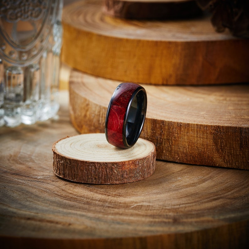May include: A dark red and black wooden ring with a black metal band. The ring is sitting on a wooden surface.