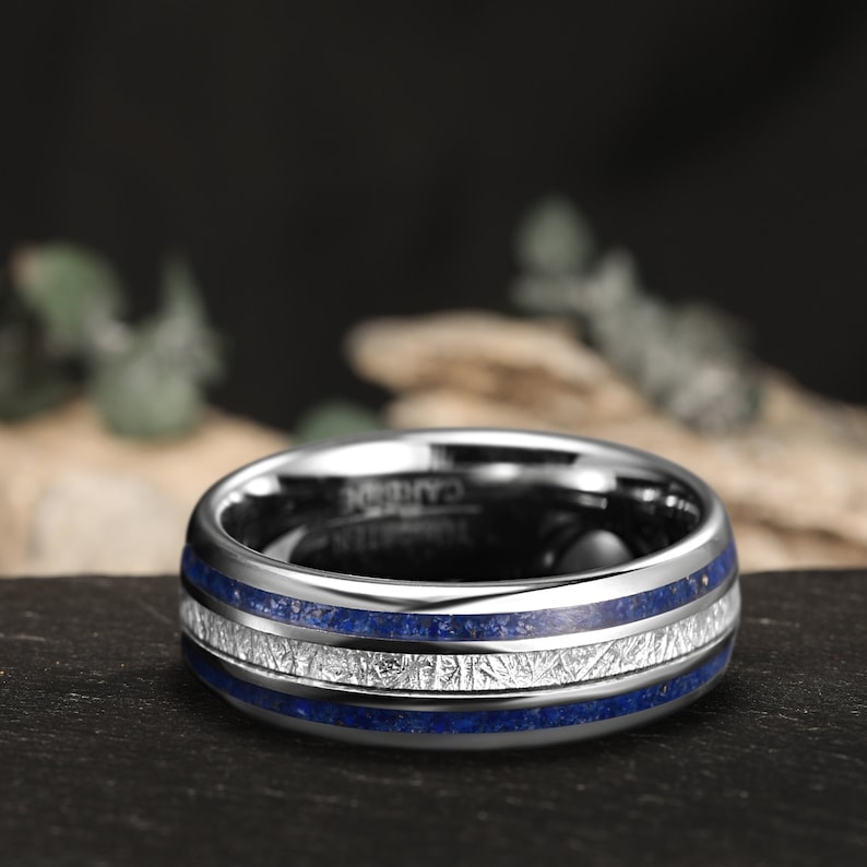 K&ouml;nnte beinhalten: Ein silberner Wolframcarbid-Ring mit zwei blauen und wei&szlig;en Einlegeb&auml;ndern. Der Ring hat eine geb&uuml;rstete Oberfl&auml;che und eine abgeschr&auml;gte Kante.