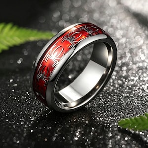 May include: A polished silver-tone metal ring featuring a wide, recessed center band with a vibrant red inlay. The red inlay is decorated with a repeating pattern of black spider silhouettes encircling the entire circumference of the ring.