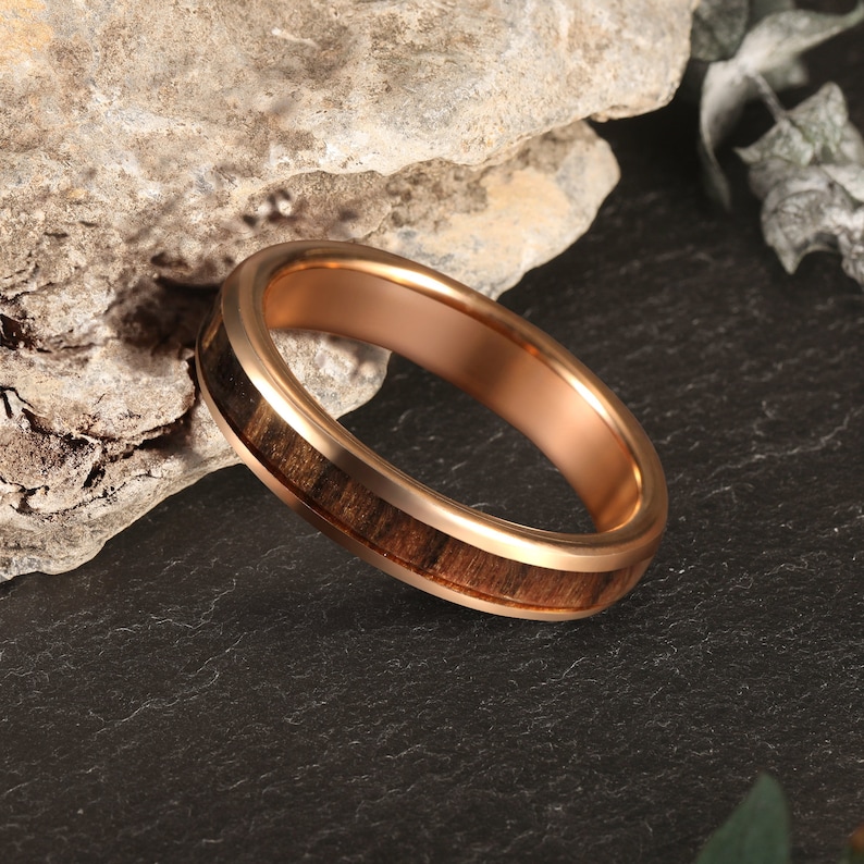 May include: A rose gold tungsten carbide ring with a brown wood inlay. The ring is on a black surface.