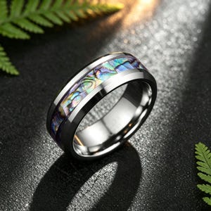 May include: A polished silver-toned metal ring featuring a wide, recessed center inlay of iridescent abalone shell. The shell displays a mosaic of vibrant blue, green, and purple hues, contrasting with the smooth, reflective metallic band edges.