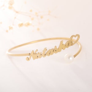 Puede incluir: Pulsera de brazalete dorada con el nombre "Natasha" grabado. La pulsera tiene un pequeño dije de perla y un recorte en forma de corazón.