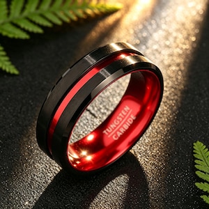Op de afbeelding: Een zwarte ring van wolfraamcarbide met een geborstelde afwerking aan de buitenkant, een dunne, felrode groef in het midden en een gepolijste rode binnenkant. De woorden TUNGSTEN CARBIDE staan in een schreefloos lettertype aan de binnenkant.