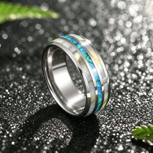 Peut inclure: Une bague en métal argenté dotée d'un large anneau avec trois canaux incrustés distincts. Le canal central contient un matériau broyé bleu vif, tandis que les deux canaux extérieurs sont remplis d'une incrustation de coquillage blanc irisé.