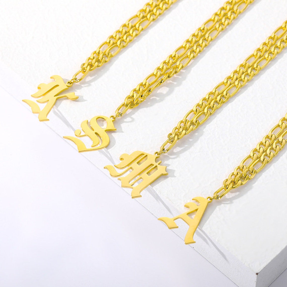 May include: Four gold chain necklaces with different letter charms. The charms are in a gothic font style. The chains are layered and have a curb link design.