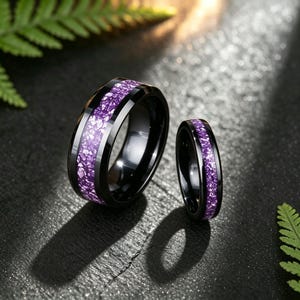 Pode incluir: Dois anéis pretos com acabamento polido, cada um apresentando uma incrustação central de material roxo triturado. O anel mais largo está posicionado à esquerda, enquanto o mais estreito está à direita, ambos exibindo uma banda escura e lisa.