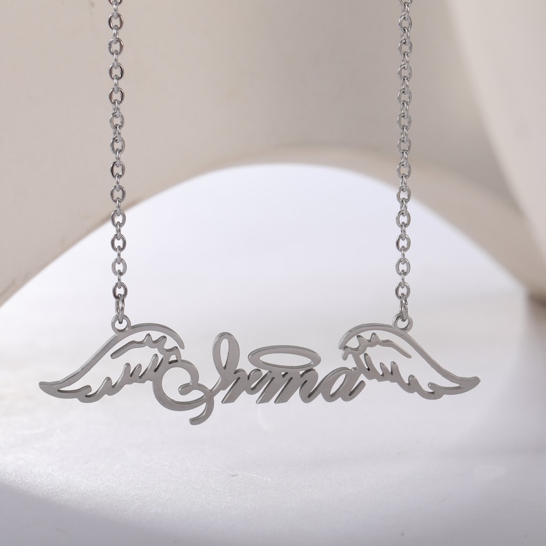 Puede incluir: Collar de plata con una placa de nombre que dice "Emma" con alas de &aacute;ngel y un halo sobre el nombre.