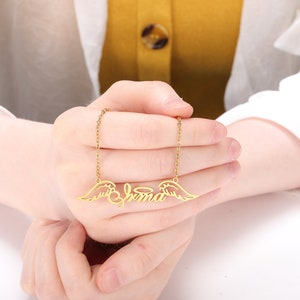 Puede incluir: Collar de oro con una placa de nombre que dice "Emma" y alas de &aacute;ngel a cada lado.