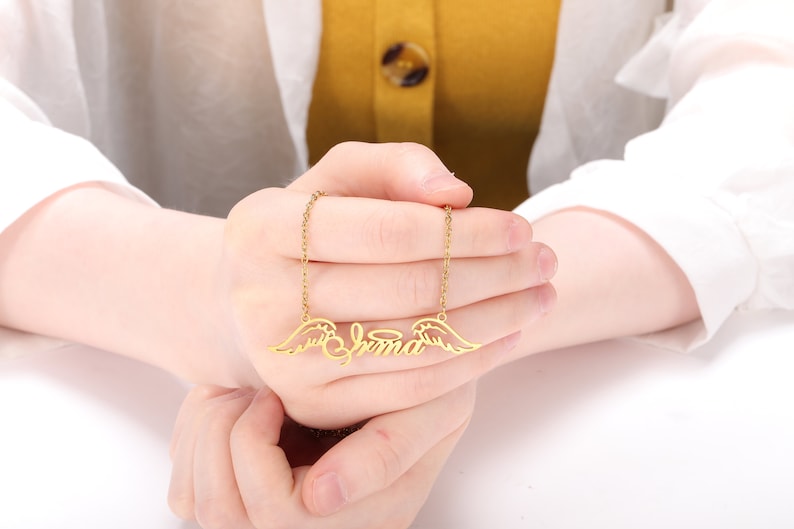 Puede incluir: Collar de oro con una placa de nombre que dice "Emma" y alas de &aacute;ngel a cada lado.