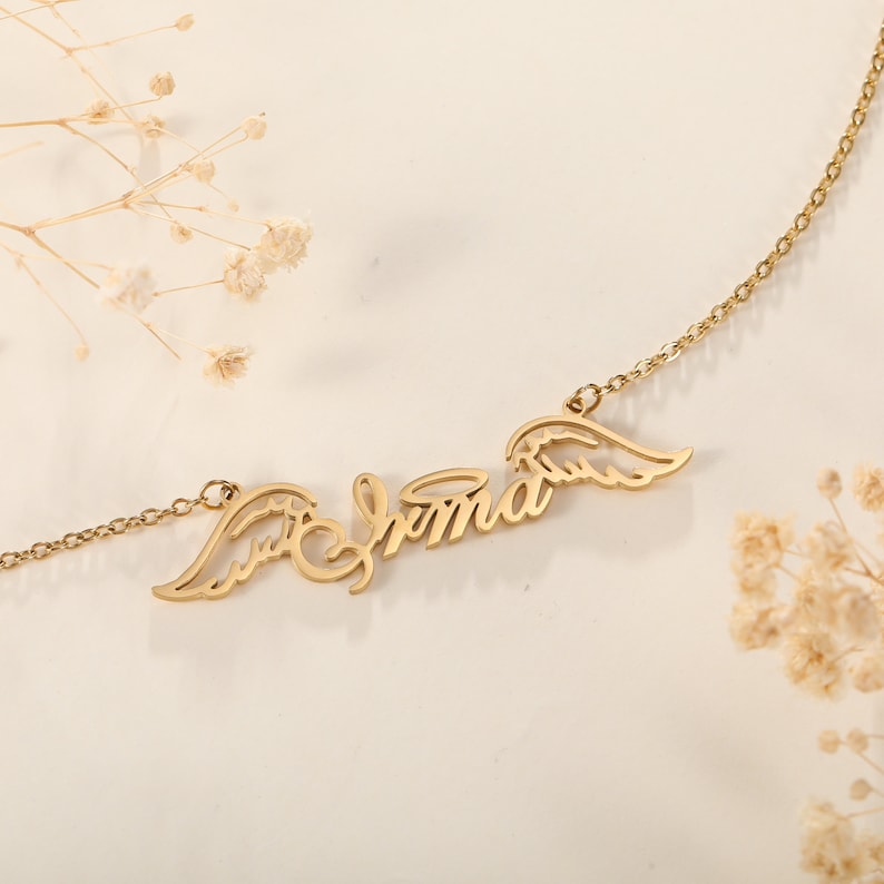 Puede incluir: Collar de oro con el nombre "Emma" escrito en cursiva con alas de &aacute;ngel a cada lado.