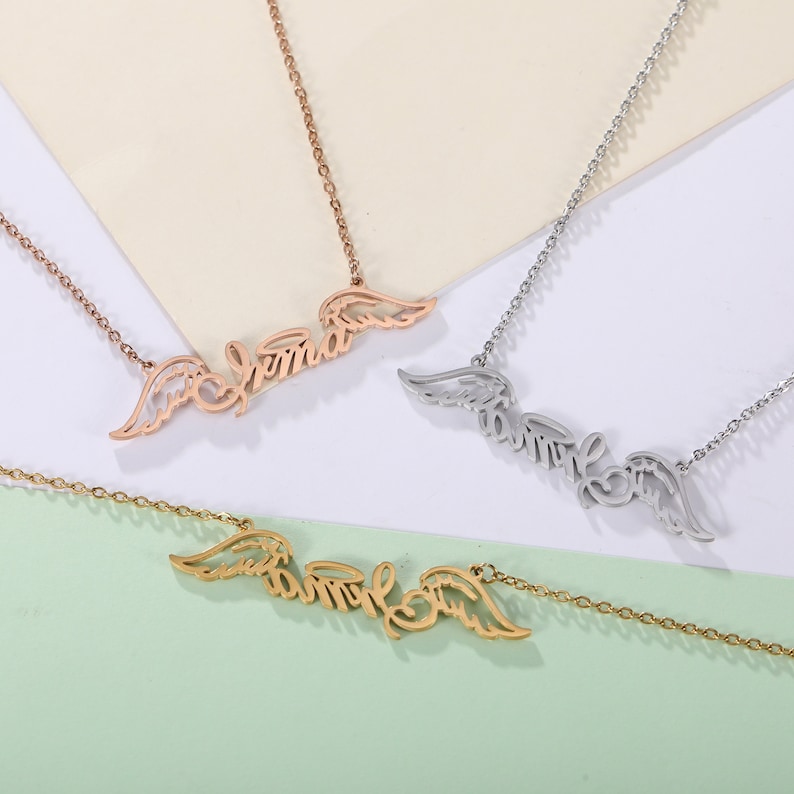 Puede incluir: Tres collares de oro con alas de &aacute;ngel y un nombre inscrito en cada uno. Los nombres son "Emma", "Emily" y "Angel".